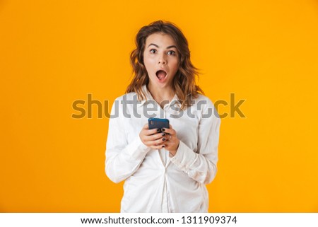 Portrait of a cheerful young woman wearing white shirt standing isolated over yellow background, holding mobile phone