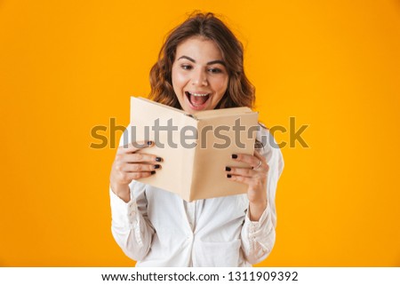 Portrait of a cheerful young woman wearing white shirt standing isolated over yellow background, holding a book