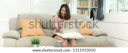 Attractive young asian woman shopping online by using notebook computer , paying by debit or credit card, sitting on sofa in beautiful decoration living room, happy lifestyle with technology concept.