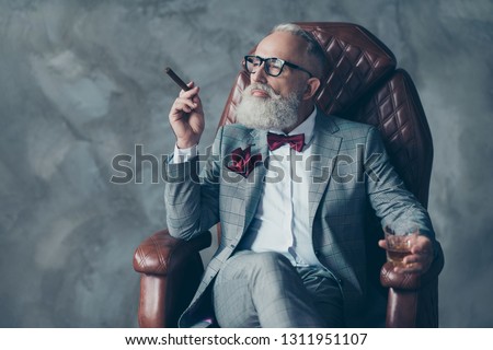 Portrait of posh chic classy virile dreamy trendy dreaming groomed brutal big company chief millionaire sitting on leather armchair smoking cigarette drinking beverage isolated on grey background Royalty-Free Stock Photo #1311951107