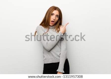 Redhead girl over white wall surprised and pointing side