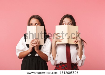 Portrait of funny girls in school uniform reading books while standing isolated over red background