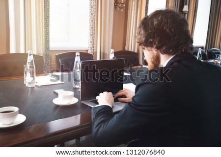 businessman in a suit sits alone in a conference room at a table with a laptop back.