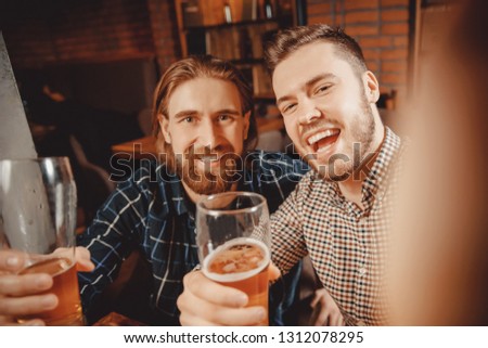 Selfie time. Two bearded hipster friends relax bar, take pictures on phone, drink beer and smile widely. They have fun. Photo for memory in social networks