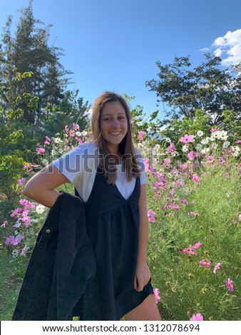 Happy, fun and beautiful young fashionable woman wearing a simple black dress with a white shirt posing in a colorful park with soft pink flowers during the spring, summertime outdoors