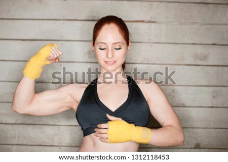 Young woman in a black sports bra and yellow boxing hand wraps looks down shyly as she flexes a muscular while poses for a portrait in front of a wooden wall.