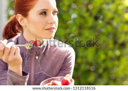 Pretty young woman sits in the sun eating a bowl of strawberries with a fork as she poses for a portrait.