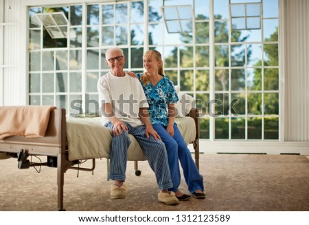 Smiling senior man being comforted by a female nurse inside his bedroom.