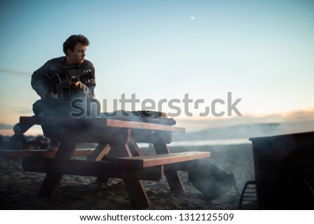 Guitar player on beach at dawn with smoke from dying campfire.