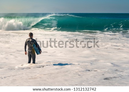 Man on the seashore prepares to surf, wears a black neoprene suit and in his hand has a blue bodyboard. the water covers him to the knee while watching a huge wave breaking
