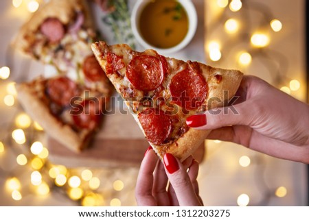 Christmas and New Year atmosphere. Womans hand takes slice of Italian pizza with melting tomato, pepperoni and cheese on a white marbel cutting board. Background with lights in bokeh and selective