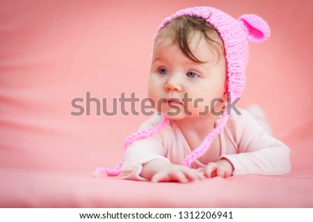Cute sweet Caucasian 6 months old baby girl in a funny pink hat with ears on a pink background.