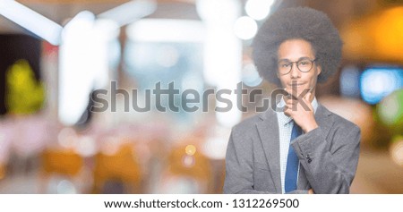 Young african american business man with afro hair wearing glasses looking confident at the camera with smile with crossed arms and hand raised on chin. Thinking positive.