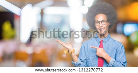 Young african american business man with afro hair wearing glasses and red tie amazed and smiling to the camera while presenting with hand and pointing with finger.