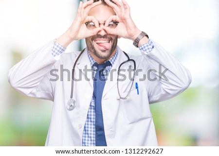 Handsome young doctor man over isolated background doing ok gesture like binoculars sticking tongue out, eyes looking through fingers. Crazy expression.