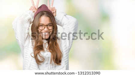 Young beautiful brunette hipster woman wearing glasses and winter hat over isolated background Posing funny and crazy with fingers on head as bunny ears, smiling cheerful
