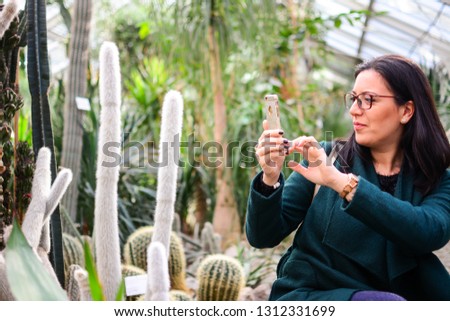 A beautiful woman enjoys visiting a diverse plant world in a beautiful botanical garden. Various cactus in a glass greenhouse for protection. Taking a picture of of interesting cactus species  - Image
