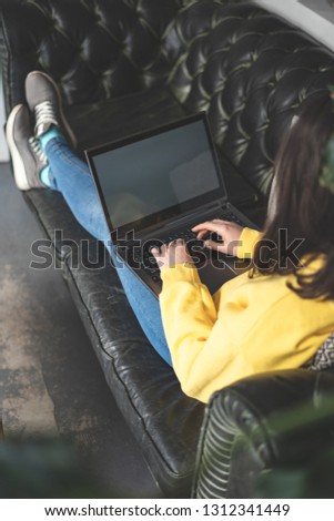 Vertical cropped photo of unrecognizable lady working on laptop inside cafe. She lying on comfort couch or sofa and writing letter to her friends