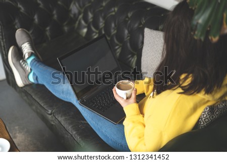 Side view cropped photo of unrecognizable lady person in stylish clothes inside cafe. She holding white mug of beverage and using her laptop lying on sofa or comfort couch