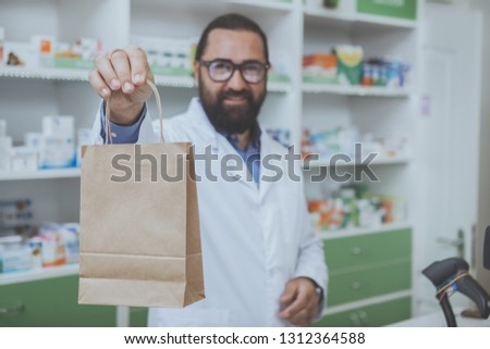 Happy handsome male pharmacist smiling joyfully holding out paper bag with medications to the camera. Attractive mature chemist working at his drustore, copy space