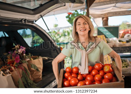 Portrait of a cheerful middle-aged woman lifting a crate of tomatoes at a farmer's market.