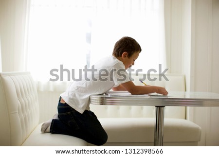 Young boy kneeling at a dining table doing his homework.