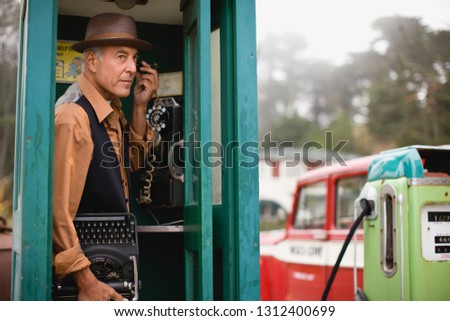 Mature man wearing a hat while listening on the phone in a public phone booth holding a typewriter.
