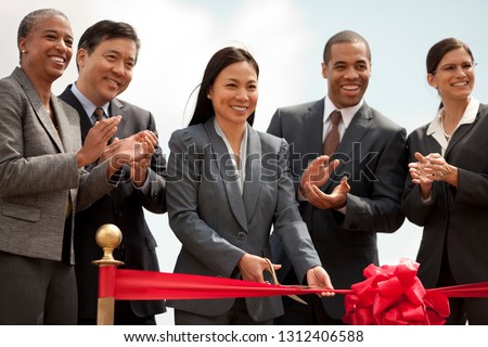 Smiling middle-aged businesswoman performs the ribbon cutting ceremony of a new development as her colleagues look on happily and applaud.