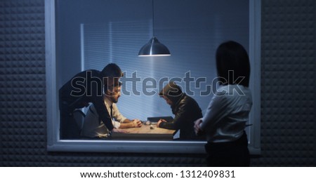 Medium shot of a male and a female investigator watching their colleague interrogating a drug dealer Royalty-Free Stock Photo #1312409831