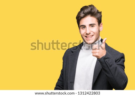 Young elegant man wearing winter coat over isolated background doing happy thumbs up gesture with hand. Approving expression looking at the camera showing success.