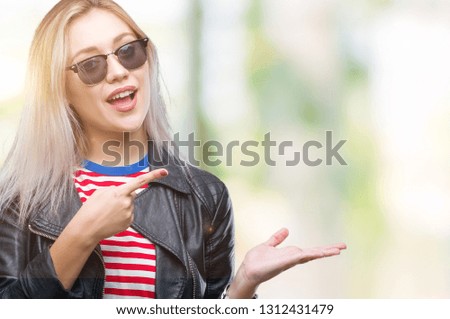 Young blonde woman wearing fashion jacket and sunglasses over isolated background amazed and smiling to the camera while presenting with hand and pointing with finger.