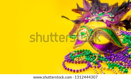 Happy Mardi Gras and Fat Tuesday carnival concept with close up on a face mask full of color, feathers and texture and golden, green and purple beads isolated on yellow background with copy space
