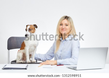 Young cute blonde caucasian girl, watches straight, holds her Jack Russell Terrier and sits by the desk in the office with the white background. Dog looks away. Laptop, cell phone and documents.