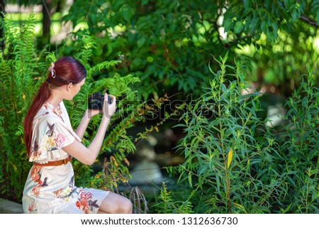 Young girl with red hair in a floral summer dress out in the garden taking photos with her camera