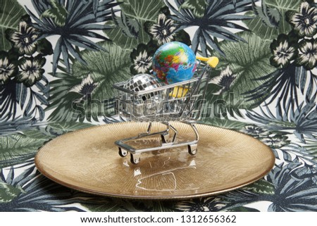 a disco ball and a planet earth in a supermarket shopping cart in a golden plate on a tropical motif background. Minimal still life color photography