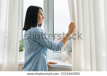 Young woman in blue shirt opening curtains looking out the window in the morning in the room, cloudy day in the city.