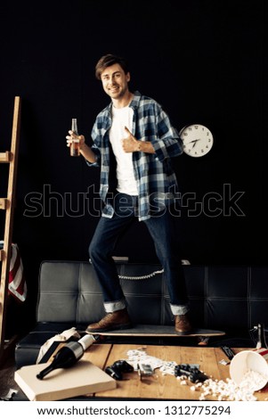 cheerful man holding bottle and showing thumb up in messy living room 