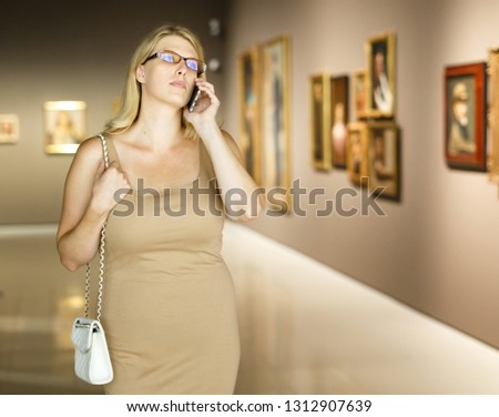 Woman visiting exposition of painting and talking on her mobile phone in art museum