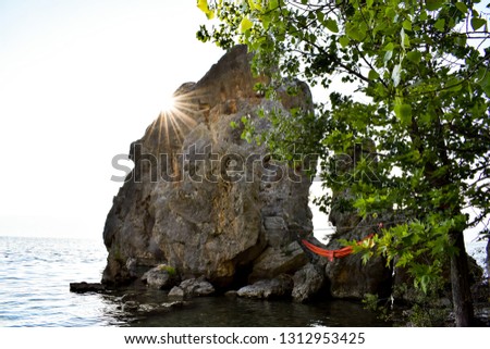 Sun over big rock on wild rocky beach on the shore of Ohrid lake. Red hammock hanging between trees and rock above water.