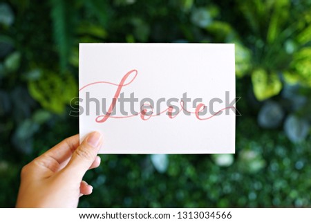 Hand holding card with modern calligraphy text "Love" on green nature background.                     