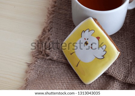 Gingerbread  with the image of a chicken is on a table. Gingerbread is made for Easter. The cup with hot tea is near gingerbread.