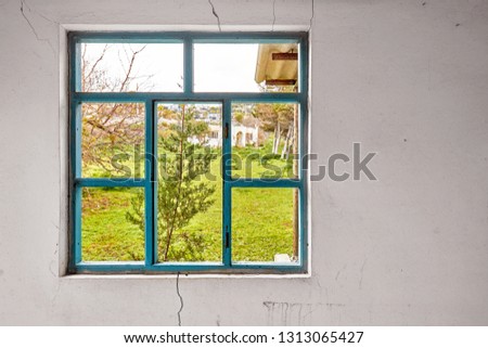 Interior of a ruined house with old, dirty and cracked white wall and a blue window frame with no glass viewing a green lawn landscape