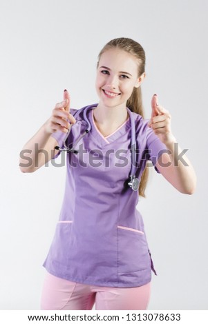 Woman doctor blonde young light gray background studio day beautiful one looks with a smile surprised rejoices thumbs up. Indicates when meeting approves the selection.