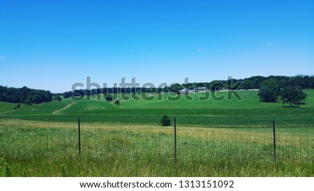 There is a beautiful green field in the southern Indiana countryside.  Bright blue sky is in the upper third of the picture, with a line of fence across the foreground.  