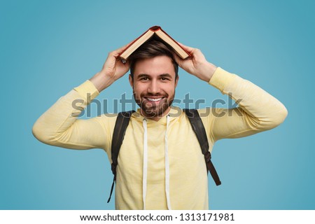 Attractive young man with backpack smiling and keeping open book on head while standing on blue background