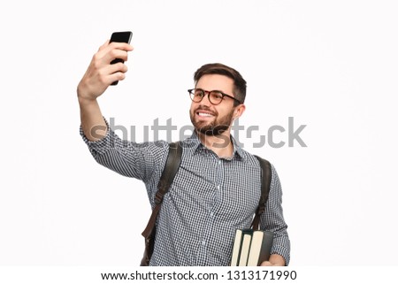 Handsome bearded student with backpack and books taking selfie using phone on white background