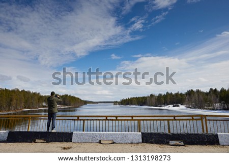 A man in a green jacket photographs nature on the phone. Located on the bridge against the blue sky and white clouds