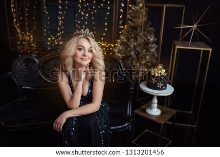 Young beautiful charismatic extraordinary girl woman in an elegant black dress in the image of the queen celebrates her birthday with a cake and candles, studio shooting, emotions, celebration, fun