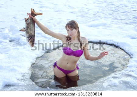 Young girl in a swimsuit standing in the ice-hole and holding a fish in her hand.