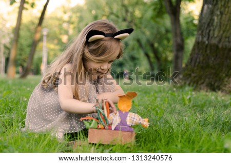 Beautiful baby girl looking for Easter egg in rabbit cartoon basket in green grass.Happy holiday traditional game for child outdoors